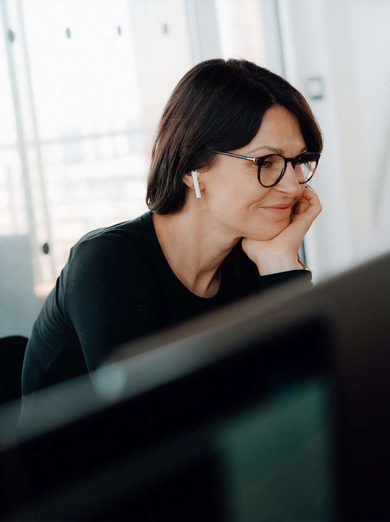 Annika arbeitet lächelnd am PC und hat das Kinn auf der linken Hand abgestützt. Sie ist von der Seite zu sehen und in ihrem rechten Ohr trägt sie einen Earpod.