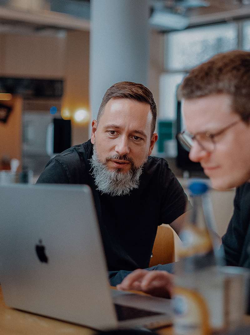 Arne und Markus sitzen gemeinsam im Co­wor­king-Space. Markus arbeitet am MacBook und Arne schaut zu.