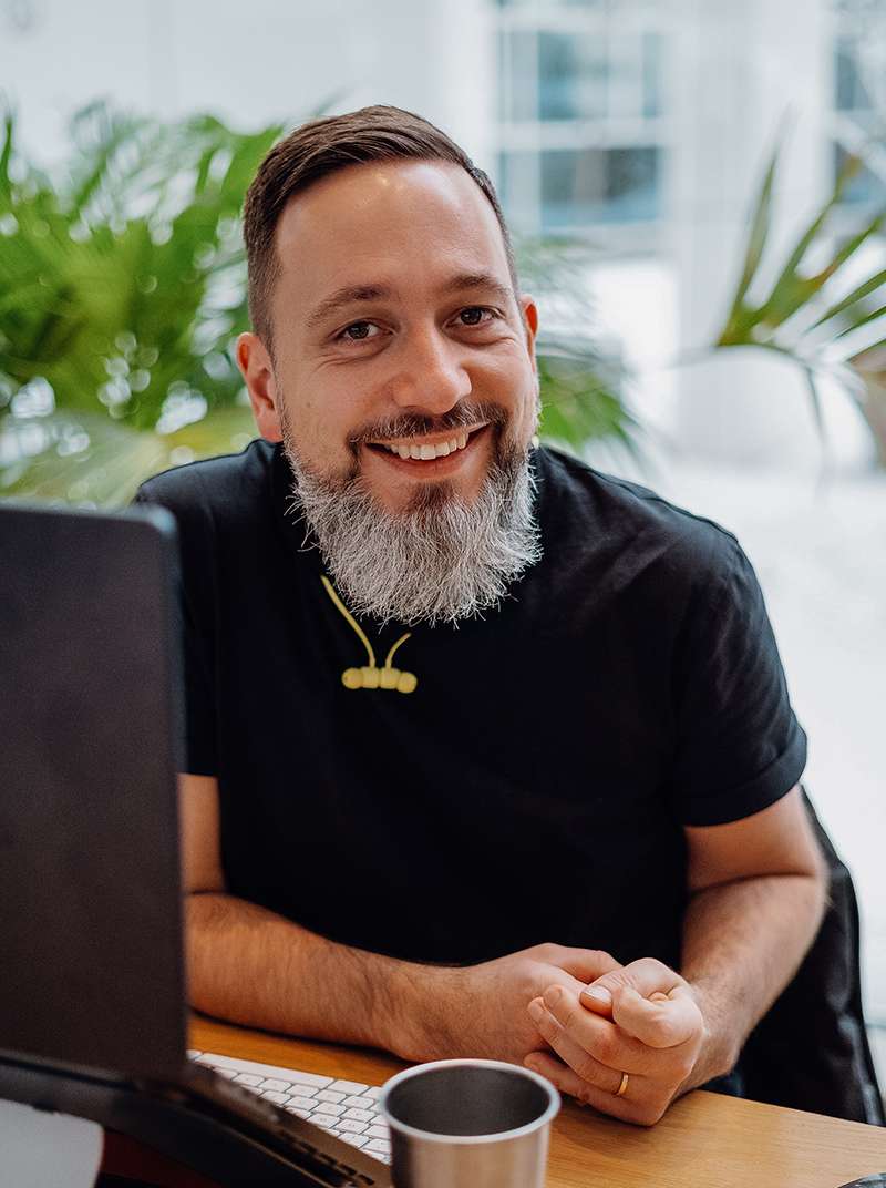 Arne sitzt grinsend an seinem MacBook im Coworking-Space und hat die Hände vor sich auf dem Tisch.