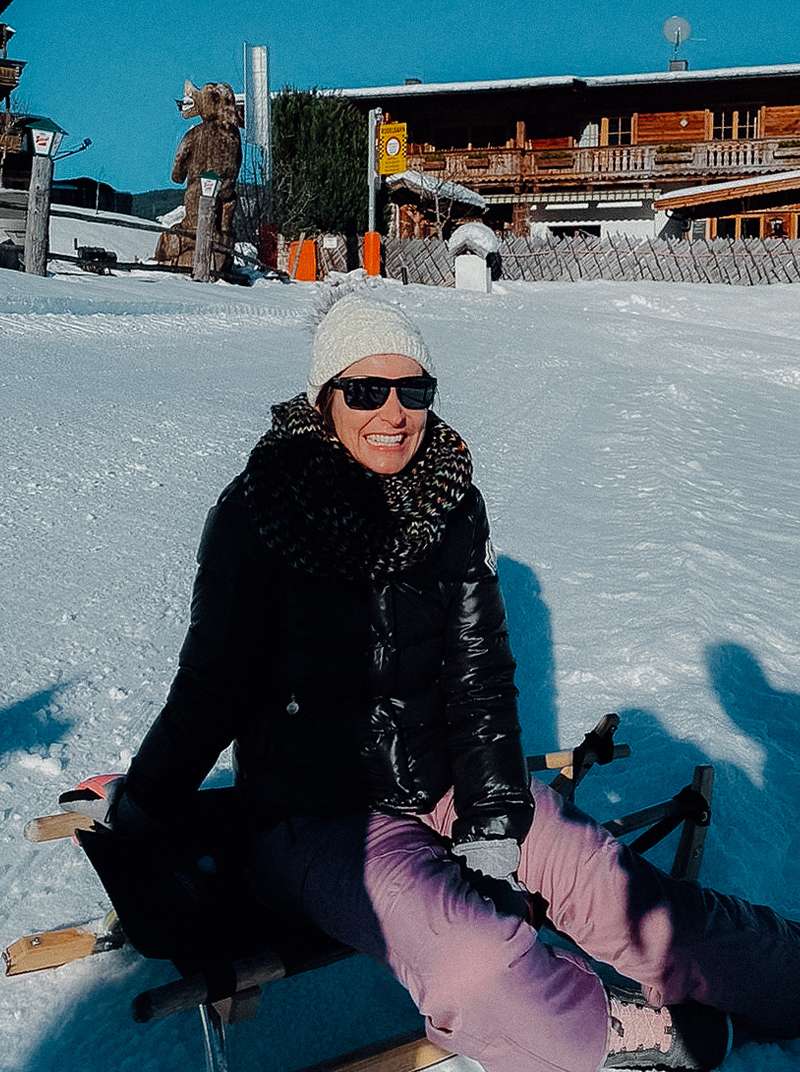 Caro sitzt bei Sonnenschein auf einem Schlitten im Schnee. Sie trägt eine lilafarbene Schneehose, schwarze Jacke, Sonnenbrille und eine weiße Pudelmütze.