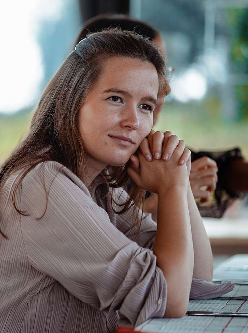 Esther sitzt an einem gedeckten Tisch und schaut verträumt am Fotografen vorbei, während sie die Hände gefaltet hat und den Kopf auf den Händen abstützt.