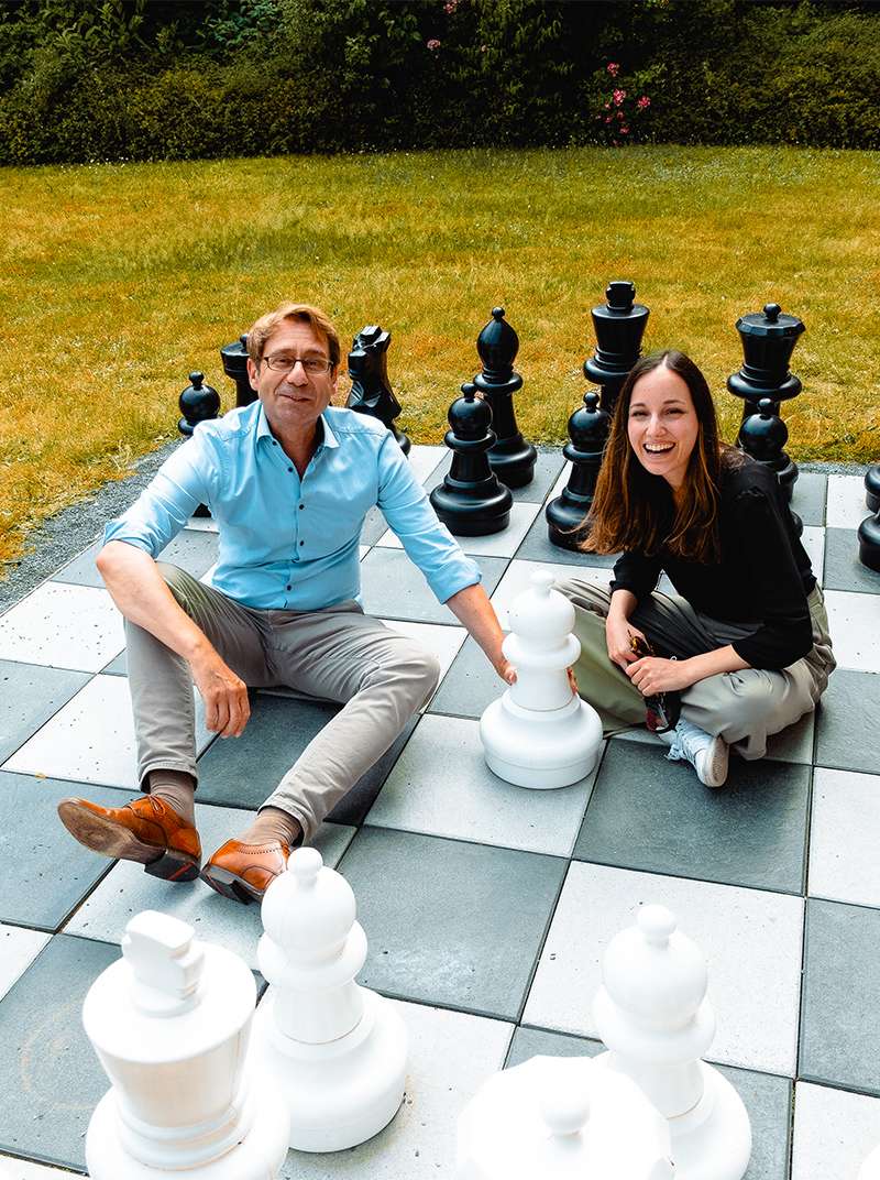 Lothar und Sarah sitzen auf einem großen Schachbrett neben schwarzen und weißen Schachfiguren. Im Hintergund sieht man eine Wiese.