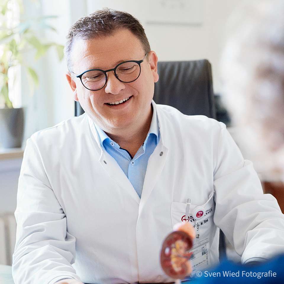 Arzt im weißen Kittel lächelt während eines Gesprächs mit einer Patientin, die man nur von hinten sieht.