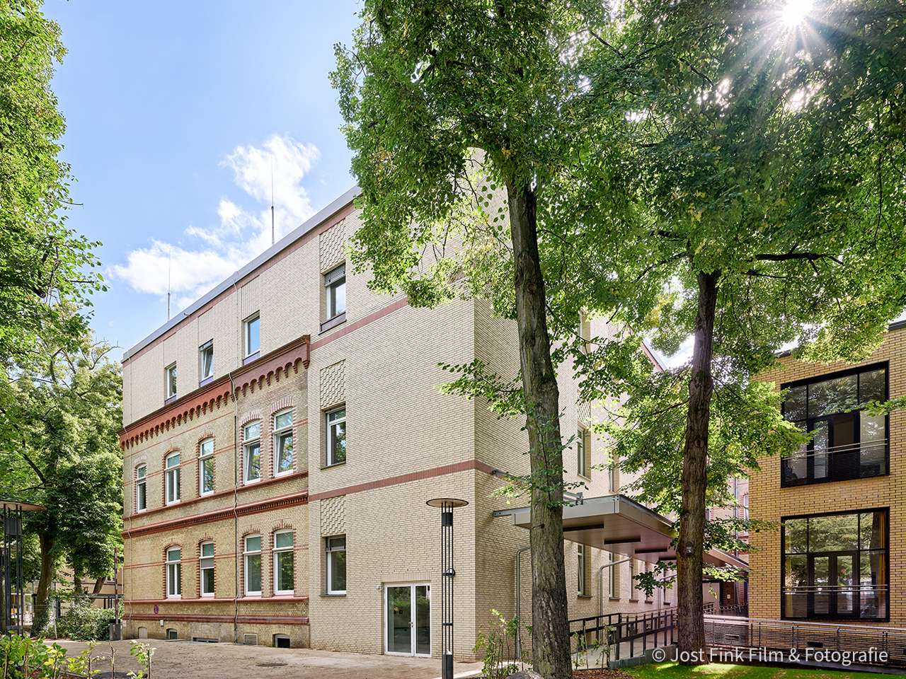 Zwei Gebäude umgeben von Bäumen unter blauem Himmel, eines mit traditioneller Fassade, das andere modern.