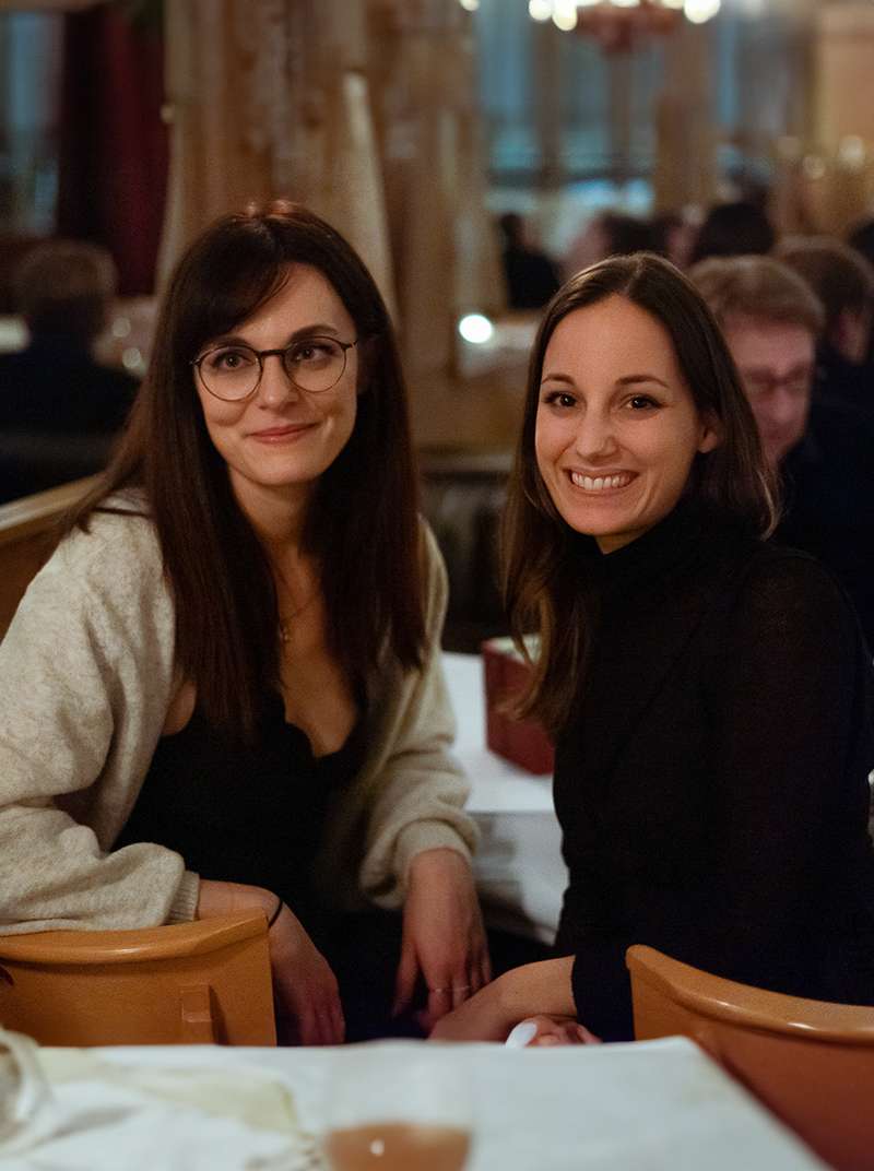 Melanie und Sarah sitzen in einem Restaurant auf Stühlen nebeneinander und schauen lächelnd über die Schulter.