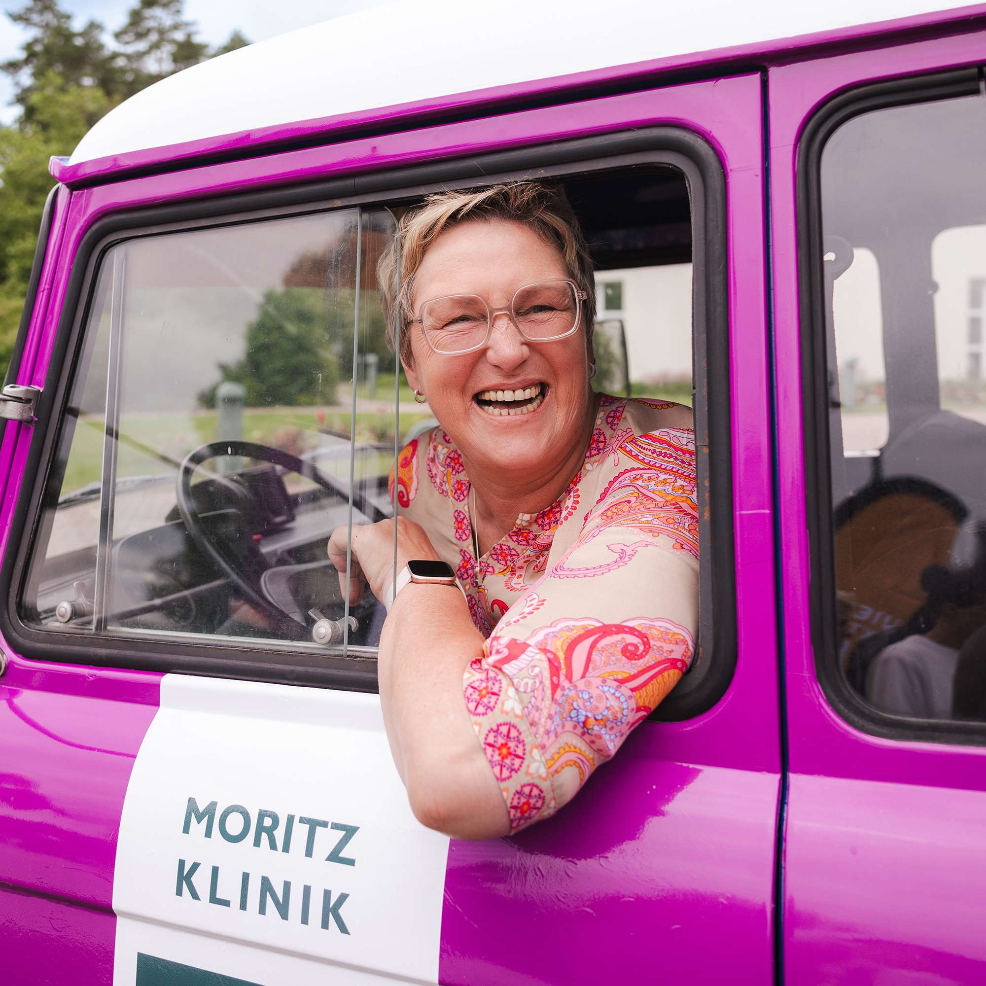Eine Frau lehnt sich aus dem Fenster der Fahrerseite eines Autos und strahlt in die Kamera.