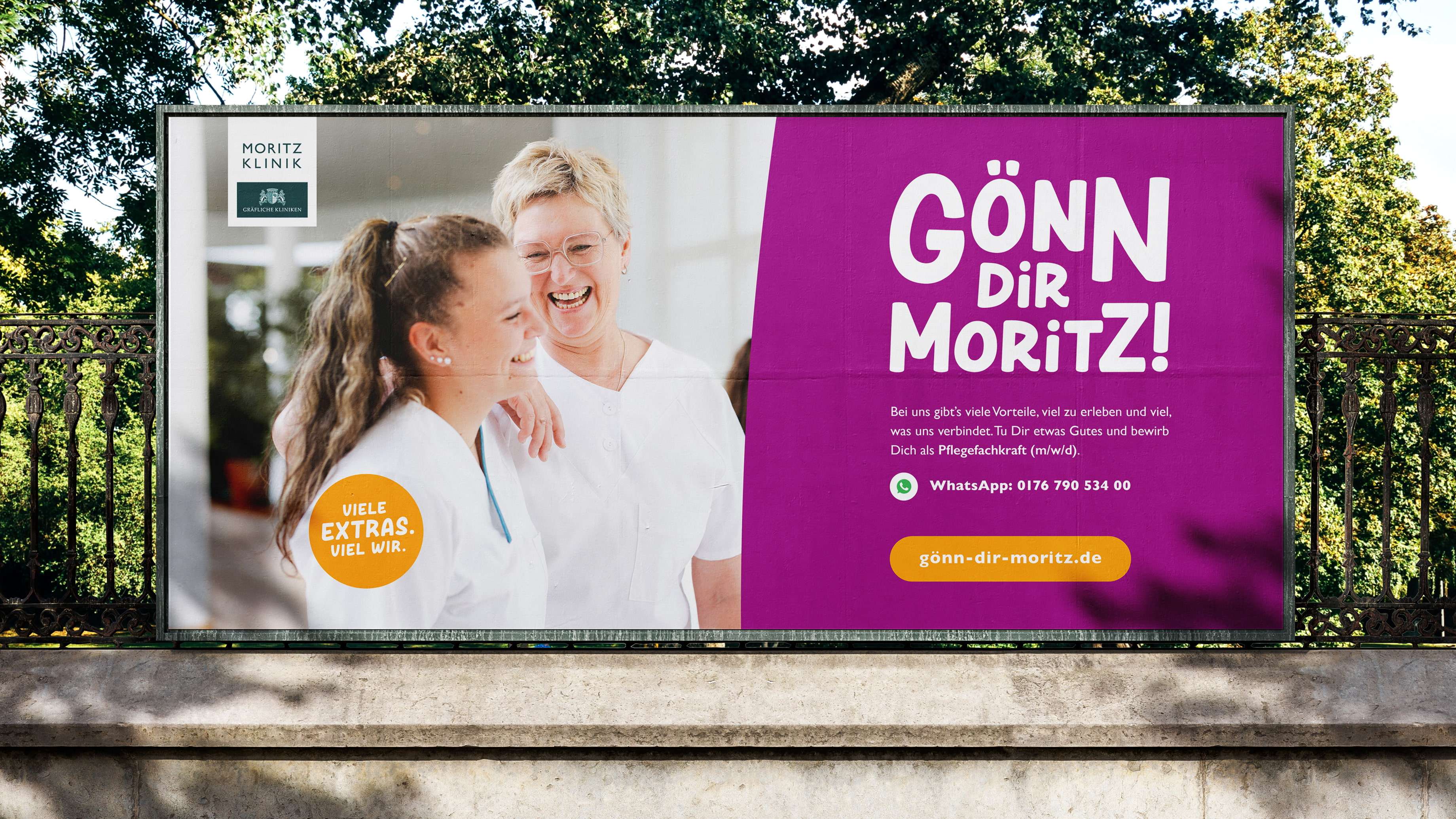 Auf einer großen Out-of-Home-Plakatwand hängt ein Plakat der Personalkampagne der Moritz Klinik. Es zeigt zwei lachende Frauen in Kasak, den Slogan der Kampagne “Gönn dir Moritz” und den Aufruf, sich als Pflegefachkraft zu bewerben.