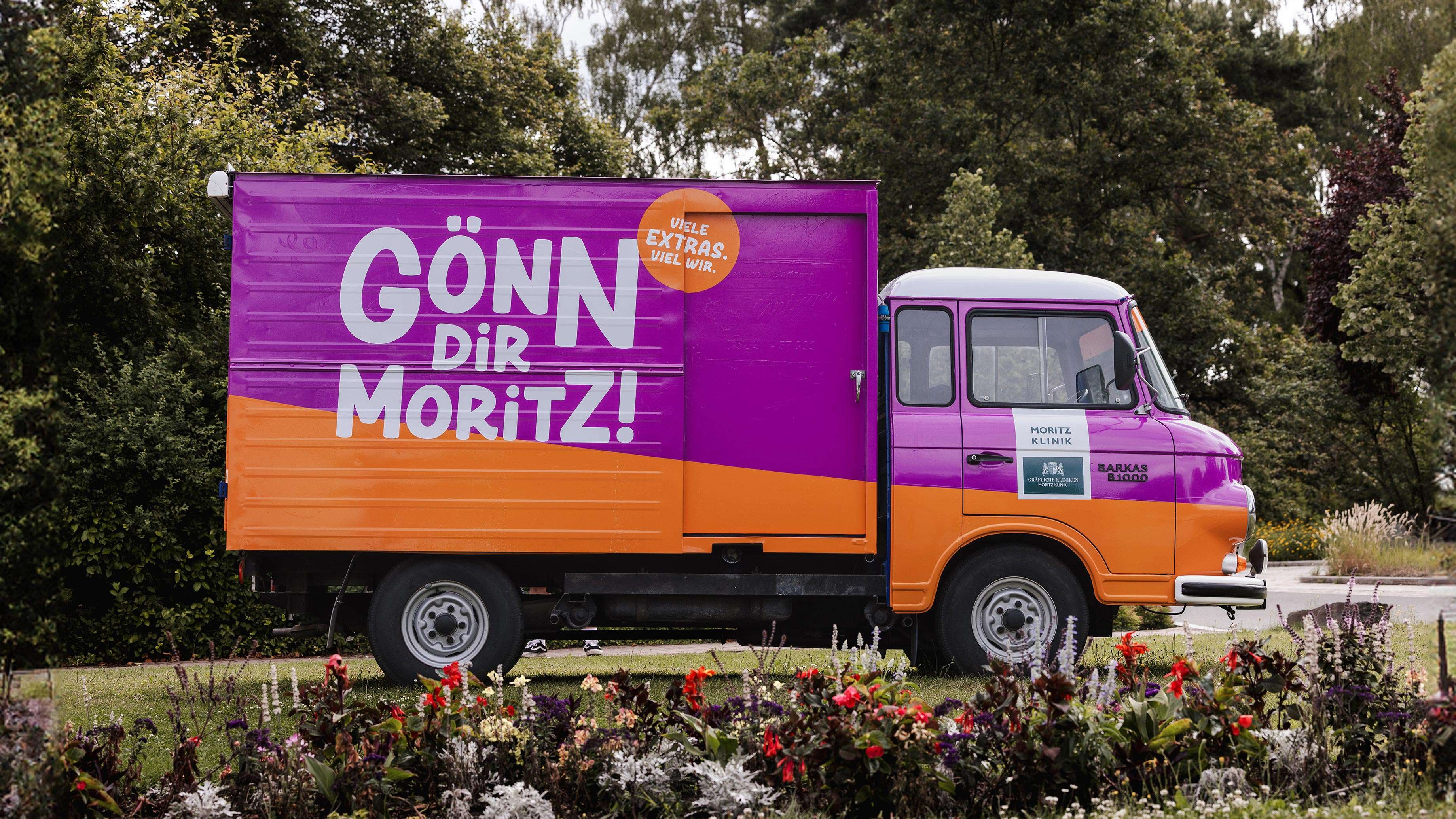 Auf einer Wiese steht ein kleiner LKW. Er ist in den Farben der Personalkampagne der Moritz Klinik, violett und orange, foliert. Auf der Seite ist der Slogan der Kampagne “Gönn dir Moritz! Viele Extras. Viel Wir.” aufgeklebt.