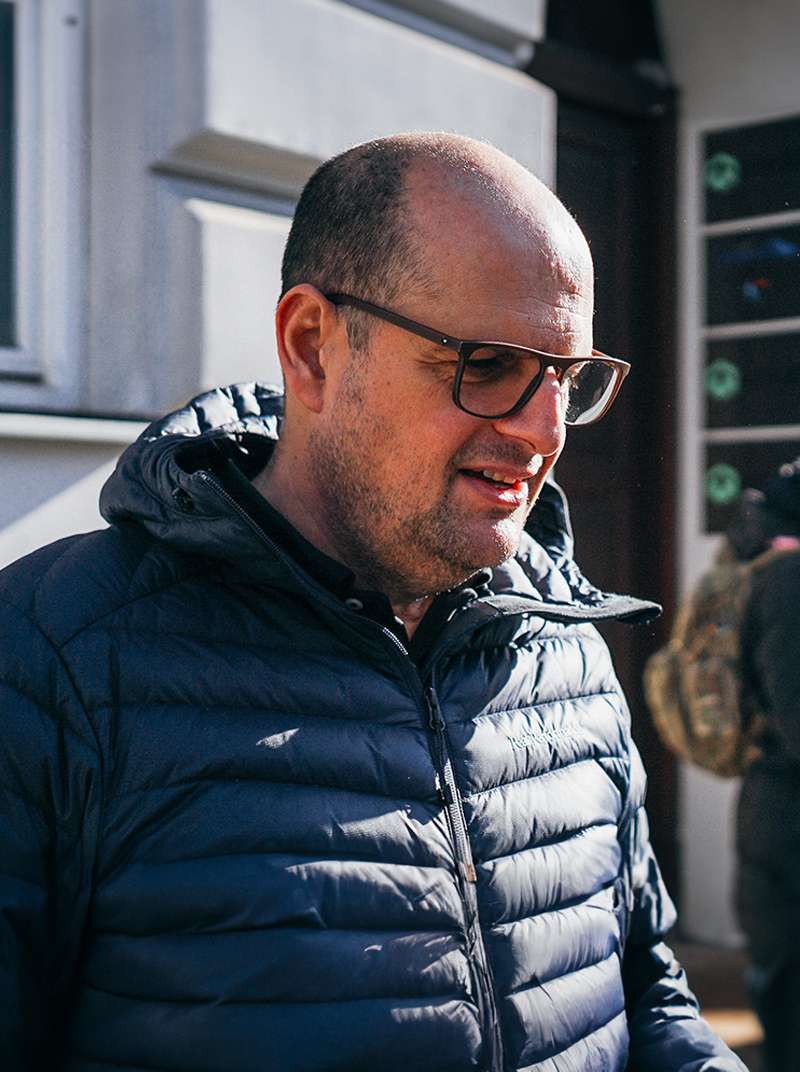 Roland steht in Winterjacke in der Sonne vor einem Gebäude und lacht.