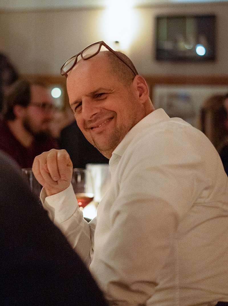 Roland sitzt breit grinsend in einem Restaurant und hat seine Brille auf den Kopf geschoben.