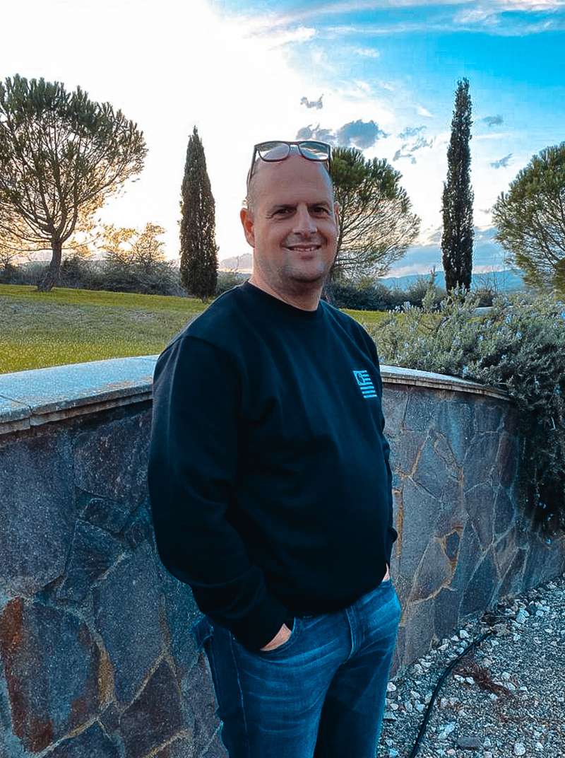 Roland steht mit seinen Händen in den Hosentaschen und Brille auf der Stirn vor einer Mauer, hinter der sich eine italienischen Landschaft mit Zypressen auftut.