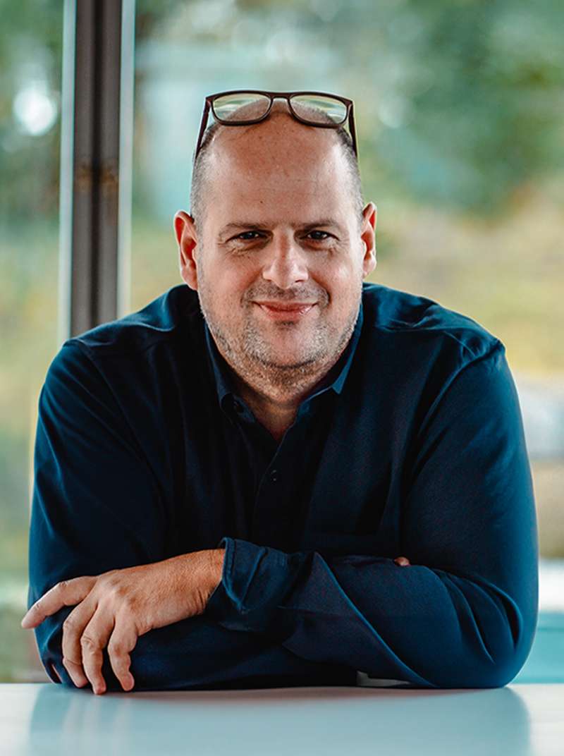 Roland steht an einem Stehtisch im Coworking-Space, er hat die Unterarme verschränkt und auf dem Tisch aufgelegt und die Brille auf den Kopf geschoben und lächelt.