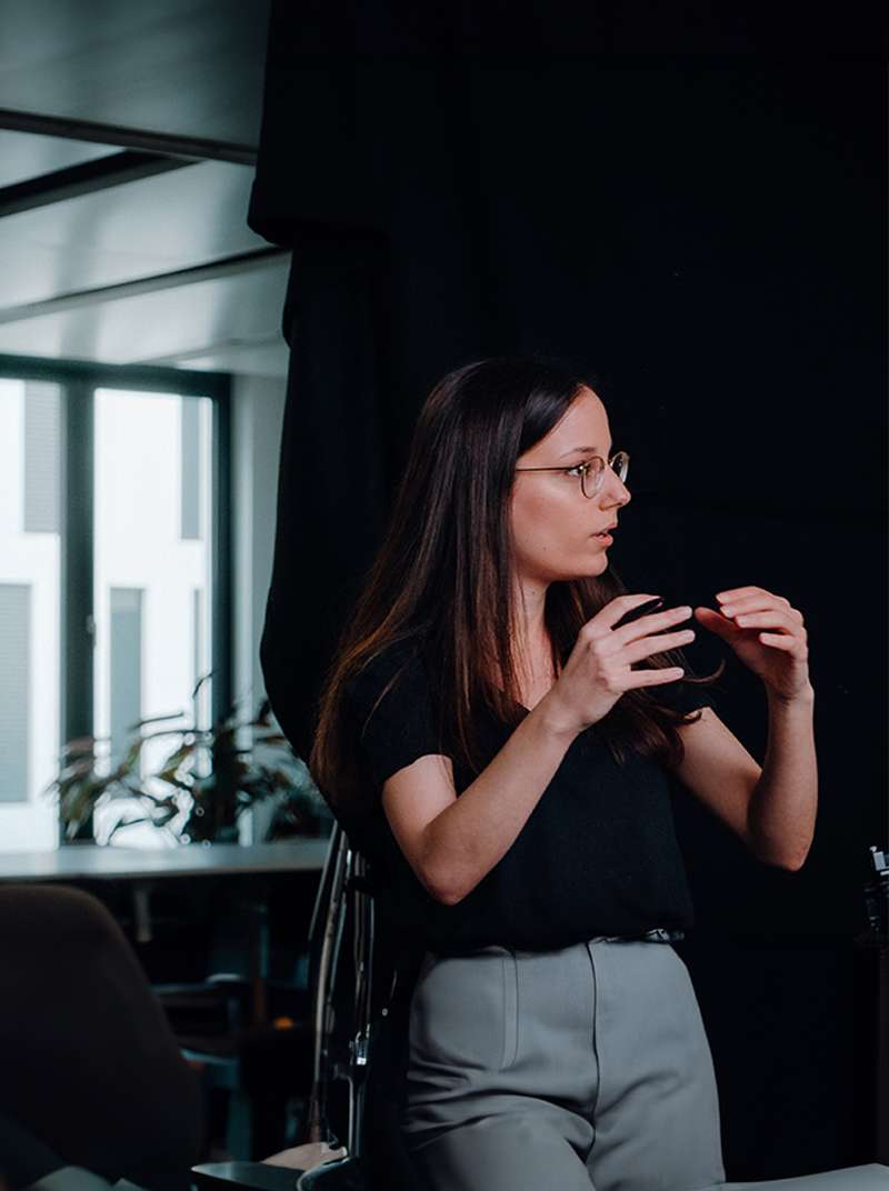 Sarah präsentiert und gestikuliert dabei mit beiden Händen vor einem schwarzen Hintergrund in einem Bürogebäude stehend.