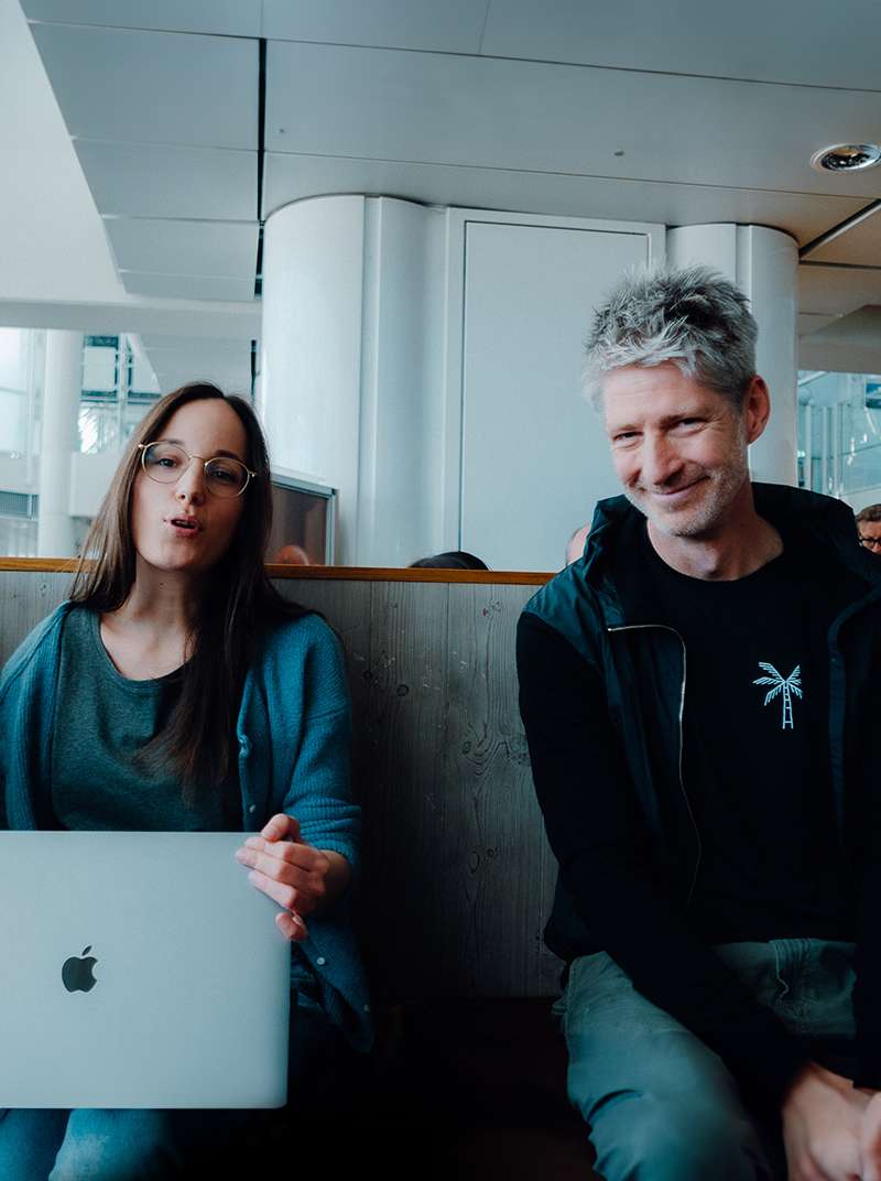 Sarah und Felix sitzen auf einer Bank. Sarah hat ein Macbook auf dem Schoß und erzählt gerade etwas, Felix lächelt in die Kamera.
