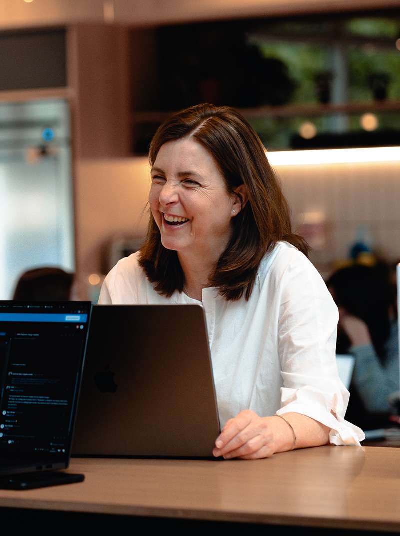 Siggi sitzt lachend hinter ihrem MacBook und hat die Hand an selbiges gelegt. Im Hintergrund ist der Coworking-Space zu sehen.