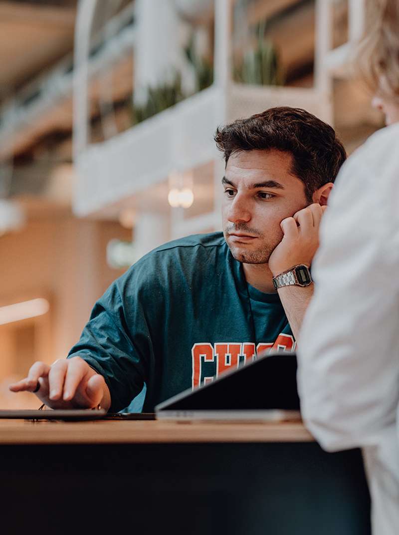 Ugur arbeitet konzentriert an einem Stehtisch im Coworking-Space, er ist von vorne zu sehen und hat den Kopf auf einer Hand abgestützt, mit der anderen Hand arbeitet er am MacBook.